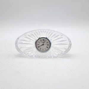 Puede incluir: Un reloj de cuarzo de cristal transparente con un bisel plateado y una esfera blanca. El reloj ovalado tiene un diseño acanalado y números romanos negros. El reloj mide aproximadamente 13 cm de largo.