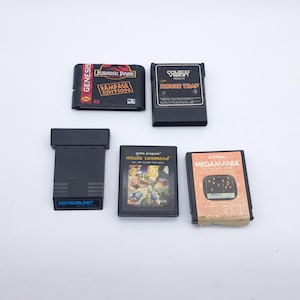 Puede incluir: Se muestran cinco cartuchos de videojuegos retro, incluyendo "Jurassic Park: Rampage Edition" y "Mouse Trap". Los cartuchos negros presentan etiquetas coloridas con títulos y gráficos de juegos clásicos.