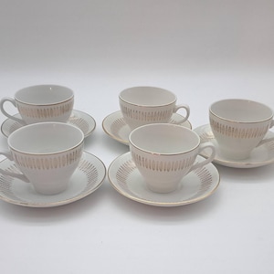 May include: A set of six white teacups and saucers. Each cup and saucer has a gold-coloured vertical stripe design. The cups have handles and are placed on matching saucers. The set is on a white background.