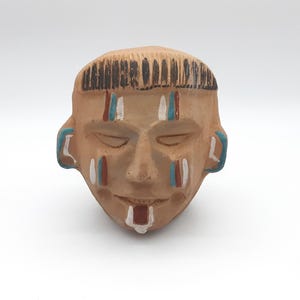 May include: A terracotta-colored ceramic mask with closed eyes and a slight smile. The mask features black hair, blue and white face paint, and red accents. The mask is a decorative item.