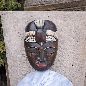 May include: A dark brown carved wooden mask with a serene face, closed eyes, and red lips. The mask has white and cream-coloured accents on the forehead and above the eyes. It is likely a decorative item, possibly for wall display.