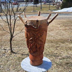 May include: A large, hand-carved wooden drum with a natural finish. The drum features intricate floral carvings and is supported by wooden struts. The drum is placed on a small, round marble-topped table. The drum's height is approximately 36 inches.