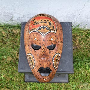May include: A carved wooden mask featuring a natural wood grain finish with black, red, and white painted accents. The mask displays stylised facial features, including almond-shaped black eyes, full black lips, and intricate geometric patterns.