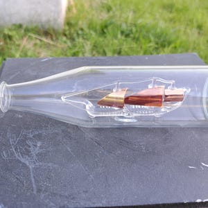 Puede incluir: Una botella de vidrio transparente con un barco en miniatura en su interior. El barco tiene velas marrones y doradas y está suspendido dentro de la botella. La botella está sobre una superficie oscura, con hierba verde al fondo.