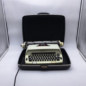 May include: A vintage off-white typewriter with black keys and a black case. The typewriter is open, revealing the keys and carriage. The case is black with a silver clasp. The typewriter has a retro design.