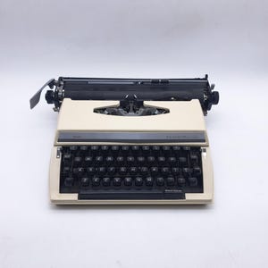 Puede incluir: Una máquina de escribir portátil de color crema con un teclado QWERTY negro. El aparato presenta un panel de control gris oscuro sobre las teclas con el texto THE ACHIEVER impreso en blanco. Incluye un carro negro y una barra espaciadora.