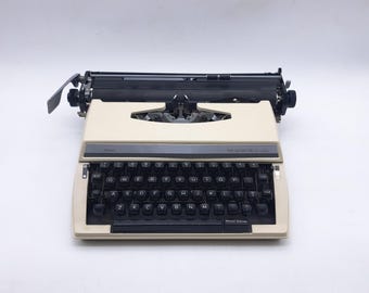 May include: A cream-colored portable typewriter with a black QWERTY keyboard. The machine features a dark gray control panel above the keys with the text THE ACHIEVER printed in white. It includes a black carriage assembly and a space bar.