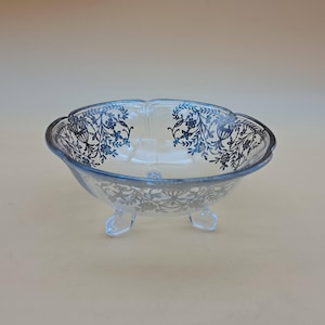 May include: A clear glass bowl with a scalloped rim and three small feet. The bowl is decorated with a blue floral pattern around the rim and on the sides. The bowl is likely used for serving or display.