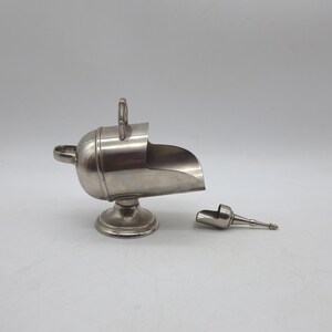 May include: A silver sugar scuttle with a scoop-shaped opening and a small handle. The scuttle sits on a round base. A separate, small silver scoop is next to it. The items are made of metal.