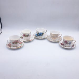 May include: Five vintage teacups and saucers are arranged on a white surface. The cups feature floral and patterned designs in various colors, including pink, blue, and gold. Each cup sits on a matching saucer, creating a classic tea set.