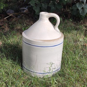 May include: A vintage stoneware jug with a handle and lid. The off-white jug features two blue stripes around the body. It is sitting on a bed of green grass.