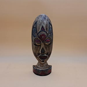 May include: A carved wooden mask with an oval shape and a detailed face. The mask features red, black, and grey accents, with a base that is also decorated with red and grey geometric patterns. The mask is a decorative item.
