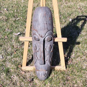 May include: A weathered, grey wooden mask with carved facial features, including large eyes, a prominent nose, and a smiling mouth. The mask is displayed on a wooden easel against a backdrop of grass.