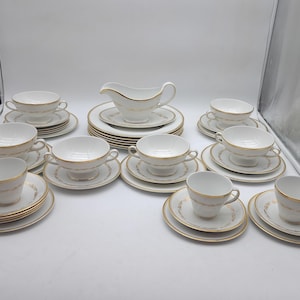 Puede incluir: Un juego de té completo de porcelana blanca con ribete dorado. El juego incluye tazas de té, platillos, boles de sopa y una salsera. El juego de té está dispuesto sobre una superficie blanca.