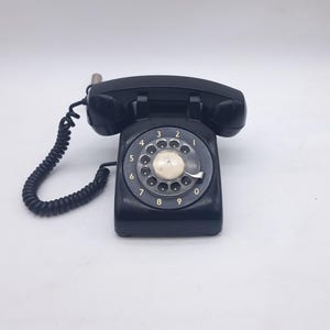 Peut inclure: Un téléphone à cadran rotatif noir vintage, avec un combiné et un cordon spiralé. Le cadran présente des chiffres blancs et un trou pour le doigt. Le téléphone est posé sur un fond blanc, mettant en valeur son design classique.