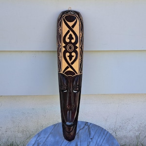 May include: A dark brown, carved wooden mask with a stylized face and intricate geometric patterns. The mask has a long, slender shape with a carved face and a decorative top section. The mask is hanging against a white wall.