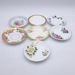 May include: A selection of seven vintage saucers, each with a unique floral pattern and gold detailing. Designs include roses, blue floral motifs, and gold rims. The saucers are displayed on a white background.
