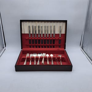 Puede incluir: Un juego de cubiertos antiguos en un estuche de madera negro con interior de terciopelo rojo. El juego incluye cuchillos, tenedores, cucharas y utensilios para servir. La cubertería tiene un diseño decorativo en los mangos.