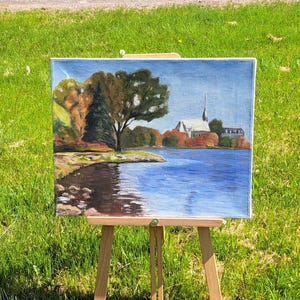 Puede incluir: Una pintura al óleo sobre lienzo que representa un paisaje sereno con un lago, árboles y una iglesia. La obra de arte se exhibe en un caballete de madera al aire libre, con hierba verde de fondo. La pintura presenta un cielo y agua azules, con árboles otoñales.
