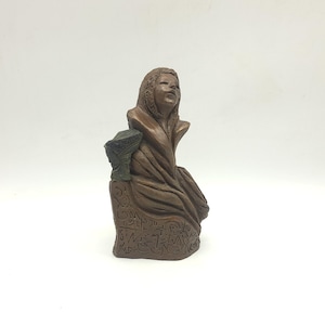 May include: A brown sculpture of a figure with a hooded head, draped in a textured garment. The base features intricate, carved symbols resembling ancient script. A small, dark, textured object is attached to the side of the figure's shoulder.