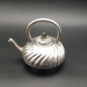 May include: A silver teapot with a curved handle and a spout. The teapot has a ribbed design and a small, round lid with a black knob. The teapot is likely made of metal.