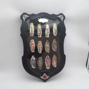 May include: A black, shield-shaped display case holding twelve decorative pocket knives. The knives have ornate handles with various designs and colors. The case has a red gem at the bottom and decorative white accents.