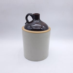 Peut inclure: Une cruche en grès vintage avec un dessus et une poignée brun foncé brillant. Le corps de la cruche est de couleur gris clair. La cruche a une forme cylindrique et une petite ouverture en haut.