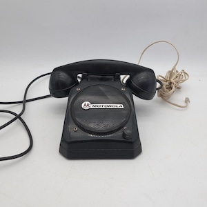 Peut inclure: Haut-parleur Motorola noir avec un combiné. Le haut-parleur a une grille en maille et le logo Motorola. Le téléphone a un cadran et un cordon. Le combiné est incurvé et a une finition noire. Le téléphone est sur fond blanc.