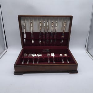 Puede incluir: Un cofre de madera para cubiertos con un acabado oscuro, abierto para revelar un juego completo de utensilios de plata. El juego incluye cuchillos, tenedores y cucharas, todos cuidadosamente dispuestos en un interior forrado de terciopelo rojo.