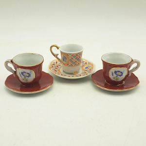 Könnte beinhalten: Drei Miniatur-Tassen und Untertassen aus Porzellan. Zwei Tassen sind dunkelrot mit Goldrand und blauem Blumenmotiv. Die dritte Tasse und Untertasse haben ein orange-blaues Gittermuster mit floralen Akzenten und Goldverzierungen an Griff und Rand.