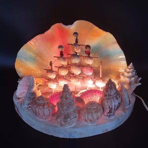 May include: A decorative lamp in the shape of a seashell. The lamp features a large, colourful shell as a backdrop, with a collection of seashells and lights. The design creates a warm glow, evoking a coastal or nautical theme.