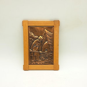 May include: A copper relief artwork in a wooden frame, showing a figure near a tent and a fire pit. The artwork depicts a person holding a long object. The frame is made of light-coloured wood. The relief is a warm copper colour.
