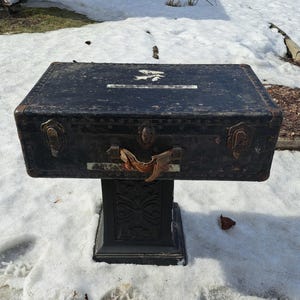 Puede incluir: Un baúl negro antiguo con herrajes metálicos y un asa, montado sobre un soporte negro decorativo. El baúl muestra signos de desgaste y envejecimiento, con una etiqueta descolorida y un aspecto desgastado. El baúl está al aire libre, sobre una superficie nevada.