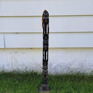 May include: A tall, dark wooden totem pole with carved faces and figures. The pole has a square base and a textured surface. A brown rope is attached to the top. The totem pole is standing on green grass.