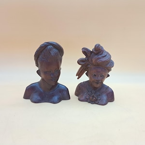 May include: Two dark brown carved wooden busts depicting figures with distinct hairstyles. The left figure has a smooth, wrapped head covering, while the right figure features an elaborate, textured updo and a floral detail on the chest area.