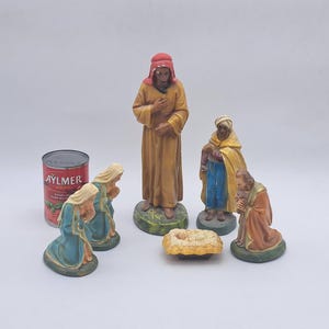 Puede incluir: Colección de figuras de belén pintadas a mano. Las figuras incluyen un hombre de pie con una túnica amarilla, figuras arrodilladas con túnicas azules y naranjas y un bebé en un pesebre. También hay una lata de tomates Aylmer.