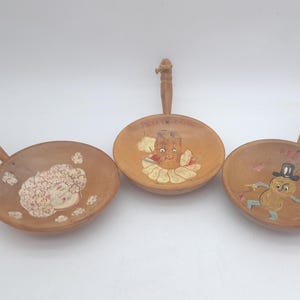 Peut inclure: Trois plats de service en bois vintage avec des poignées. Chaque plat présente un motif peint à la main unique : un visage de femme, un personnage d'arachide et un cacahuète avec un chapeau haut de forme. Les plats sont bruns avec des accents clairs.