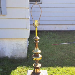 Puede incluir: Una lámpara alta de latón vintage con una base decorativa escalonada y un cuello delgado y curvado. La lámpara mide aproximadamente 122 cm de alto y se asienta sobre una base cuadrada negra. La lámpara está diseñada para sostener una pantalla.