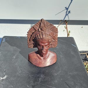 May include: A carved wooden bust of a person wearing a feathered headdress. The bust is a reddish-brown colour and has detailed carvings on the headdress and face. The bust is displayed on a dark, textured surface.