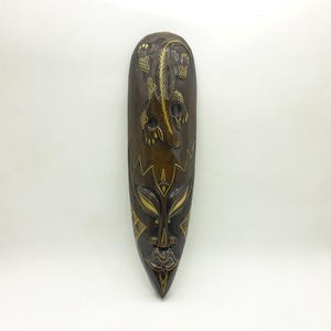 May include: A tall, narrow, dark brown wooden wall mask featuring a carved lizard at the top and a stylised face at the bottom. The mask is decorated with intricate, light-coloured etched patterns and lines that highlight the facial features and the lizard.
