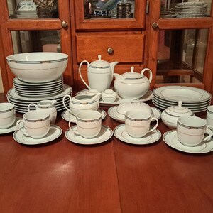 May include: A complete white porcelain tea and dinner set with a gold and teal trim. The set includes cups, saucers, plates, bowls, teapots, a sugar bowl, and a creamer. The set is arranged on a wooden table in front of a wooden cabinet.