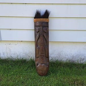 Puede incluir: Una máscara tribal de madera alta con una cara tallada y patrones intrincados. La máscara es de color marrón oscuro con una banda de material marrón claro alrededor de la parte superior y un mechón de pelo negro. La máscara está sobre hierba verde.