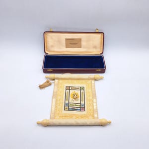 Puede incluir: Un pergamino decorativo con un fondo crema y un diseño cuadrado central con una llama y texto. El pergamino está enrollado con clavijas de madera y una borla dorada. Una caja burdeos abierta con interior azul está en el fondo.