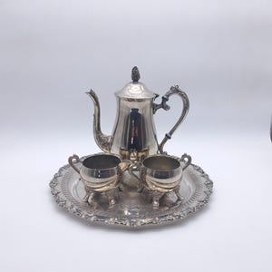 Peut inclure: Un service à thé en argent comprenant une théière, un sucrier, une crémière et un plateau de service. La théière a une poignée décorative et un bouton sur le couvercle. Le plateau et les pièces de service ont des détails ornés.