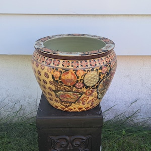 May include: A large, round ceramic planter with a wide opening. The planter is decorated with intricate floral and geometric patterns in gold, red, and black. It sits on a dark wooden stand, showcasing its ornate design.