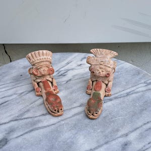 Puede incluir: Dos figuras de terracota que presentan una figura central con un tocado elaborado sentada detrás de una forma animal alargada. Las figuras están hechas de arcilla marrón claro con acentos de pintura roja y verde en los cuerpos.