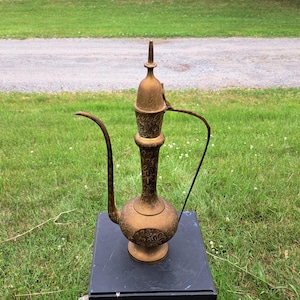 Puede incluir: Una jarra de latón vintage con un diseño ornamentado. La jarra tiene un pico largo y curvo y un asa. El cuerpo de la jarra está decorado con patrones florales. Está sobre una superficie negra, con un fondo de hierba verde y un camino de grava.