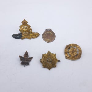 Puede incluir: Una colección de insignias y distintivos militares antiguos. Las insignias son de varias formas y tamaños, con tonos dorados y bronce. Una insignia presenta un cañón, otra una hoja de arce.