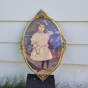 Puede incluir: Retrato ovalado antiguo de una niña en un marco dorado ornamentado. La niña lleva un vestido rosa claro y está de pie frente a un edificio. El marco tiene detalles intrincados y una estética vintage.