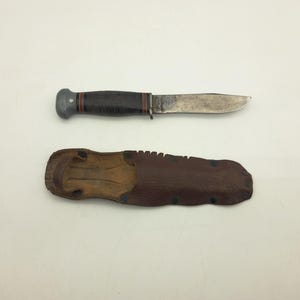 May include: A fixed-blade knife with a dark brown stacked leather handle, a silver-coloured metal pommel, and a steel blade. Below the knife is a brown leather sheath featuring a textured surface, metal rivets, and a belt loop attachment.
